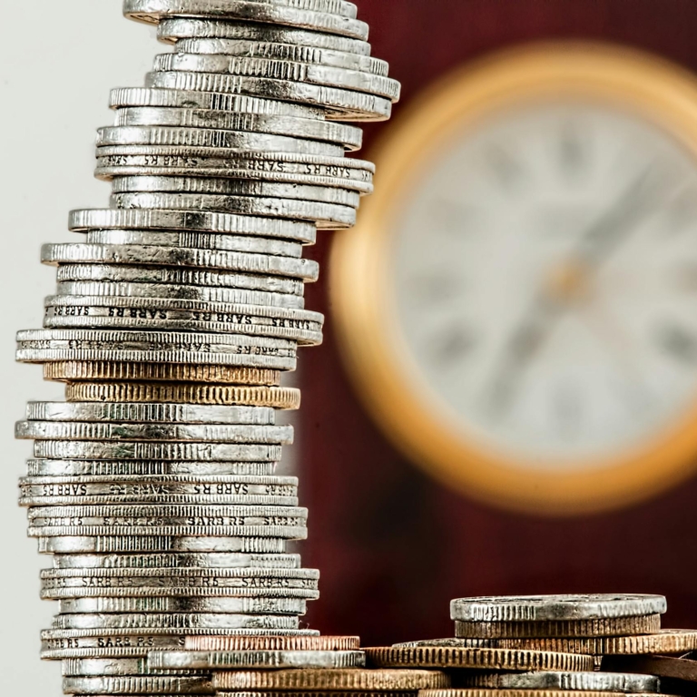 A close-up image of stacked coins with a blurred clock, symbolizing time and money relationship.